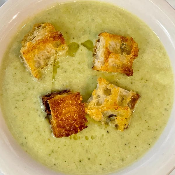 Creamy green soup with four golden-brown croutons and a drizzle of olive oil in a white bowl.