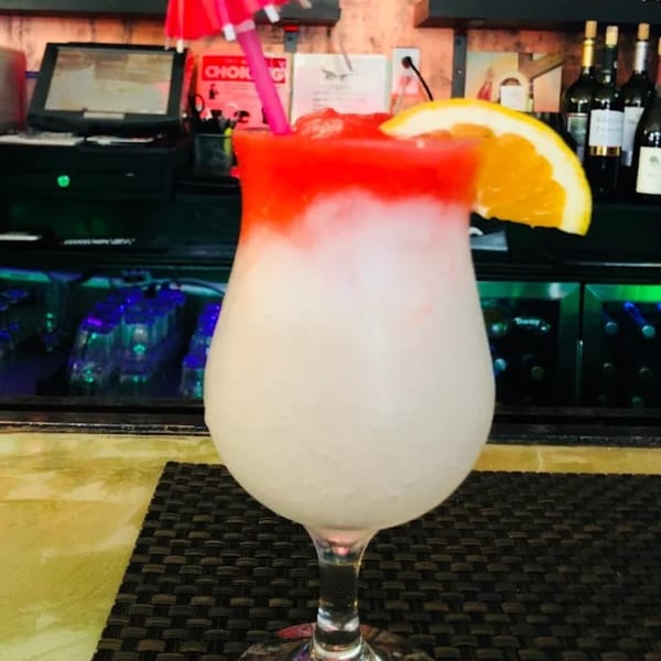 A red and white frozen cocktail with an orange slice and pink umbrella sits on a bar.