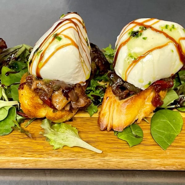 Two bruschetta pieces topped with burrata, caramelized onions, balsamic glaze, and fresh greens.