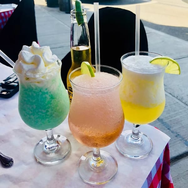 Three colorful frozen drinks, one topped with whipped cream, and a beer bottle on a table.